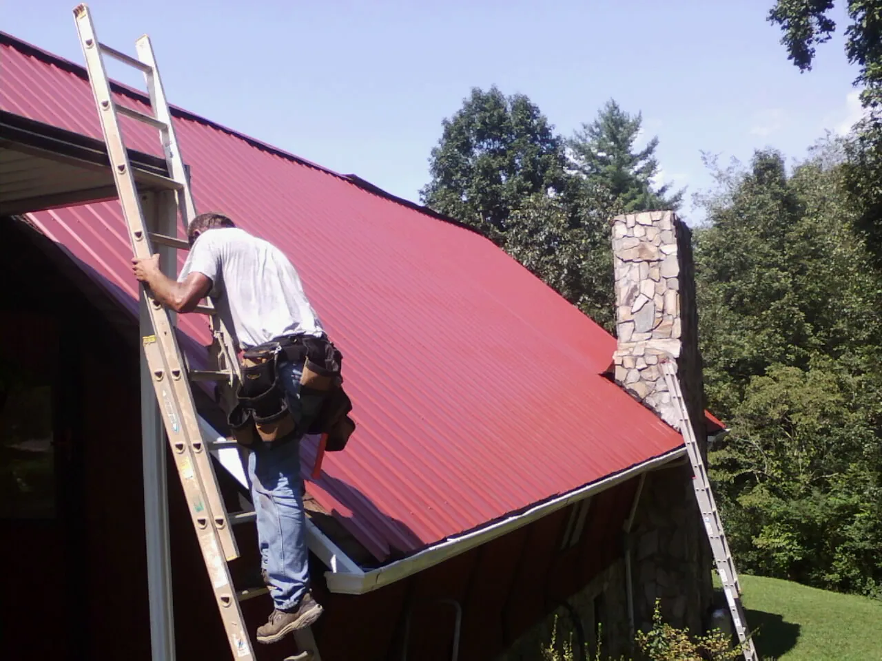 Roofer on a ladder beside a red metal roof and stone chimney — Soffit & Fascia Repair by Roofing Brothers in Jerusalem